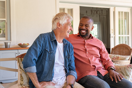 Two friends talking while sitting on couch in the courtyard. Senior man and african guy laughing while in conversation sitting outside home. Two men talking to each other and enjoying.の写真素材
