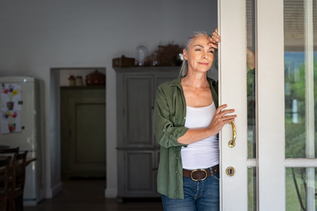 Thoughtful senior woman standing at home entrance leaning against door. Mature woman waiting husband at doorway and looking away. Lady in casual clothing standing at door and thinking.の写真素材