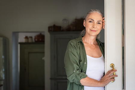 Portrait of senior woman standing while leaning against door at home and looking at camera. Smiling mature woman standing at doorway and looking at camera. Portrait of old lady relaxing and thinking with copy space.の写真素材