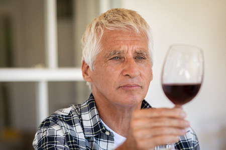 Senior man holding and looking through a glass of red wine. Old man tasting red wine at home. Retired man inspecting quality of wine during wine tasting, quality control.の写真素材