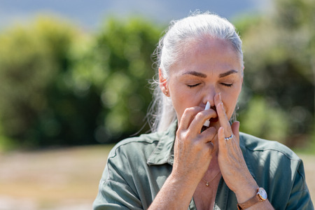 Senior woman having flu using nasal spray to help herself. Woman using a nasal spray at park for allergy. Ill mature woman with grey hair inhaling medicine against allergy.の写真素材