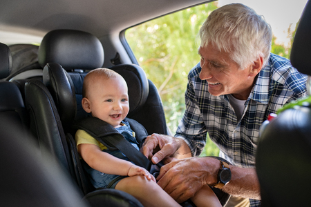 Grandfather tying baby in child seat. Cute little boy going for road trip with grandfather. Happy senior man and happpy smiling grandchild enjoying car trip. Toddler boy buckled into car seat.の写真素材