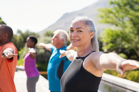 Portrait of happy senior woman practicing yoga outdoor with fitness class. Beautiful mature woman stretching her arms and looking at camera outdoor. Portrait of smiling serene ladyÂ with outstretched arms at park.の写真素材