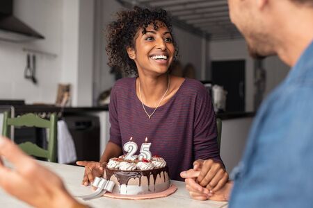 Multiethnic couple sitting at table and celebrating birthday at home in living room. Young african girl surprised on seeing 25th birthday cake with her boyfriend. Excited wife celebrating birthday with husband at home.の写真素材