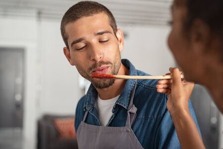 Closeup face of young man blowing on spoon to taste tomato sauce while woman cooking in kitchen. Guy in apron tasting food for dinner. Girl offering to her boyfriend a wooden spoon with sauce to taste.の写真素材