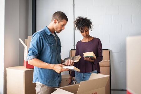 Multiethnic couple reading books while unpacking stuff after relocating to a new flat. Young man and african woman packing cardboard boxes with books. Guy and girl moving house.の写真素材