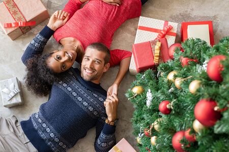 Young man and african woman lying on floor near christmas tree with presents. Top view of happy multiethnic couple lying down on floor near xmas gifts holding hands and looking at camera. High angle view of smiling couple with christmas sweater at home.の写真素材