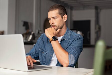 Serious young man working on laptop at home. Focused entrepreneur working on computer sitting at table. Thoughtful worried guy reading important articles and working on computer.の写真素材