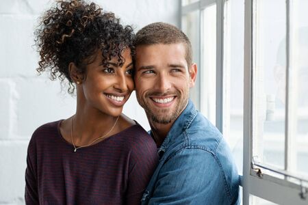 Loving multiethnic couple embracing and sitting near window. Happy girlfriend and smiling boyfriend looking away and thinking about their future together. Young man and african woman contemplating.の写真素材