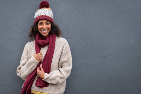 Beautiful young black woman in winter clothing standing and leaning against grey wall. Portrait of attractive african woman wearing woolen cap and scarf and looking at camera. Smiling girl feeling cold on gray background.の写真素材