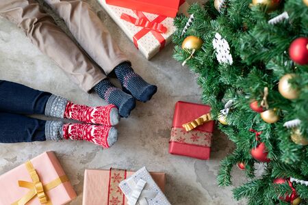High angle view of loving couple feet near christmas tree with gifts. Top view of man and woman wearing knitted warm socks relaxing at home. Closeup of legs of couple lying on floor and wearing christmas theme socks at home.の写真素材