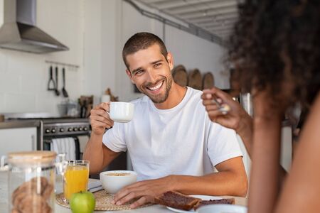 Young  smiling man and african woman having breakfast together with cup of coffee at home. Laughing multiethnic couple in conversation while eating milk and cereals . Smiling guy drinking cappuccino while having breakfast with his girlfriend in the morning.の写真素材