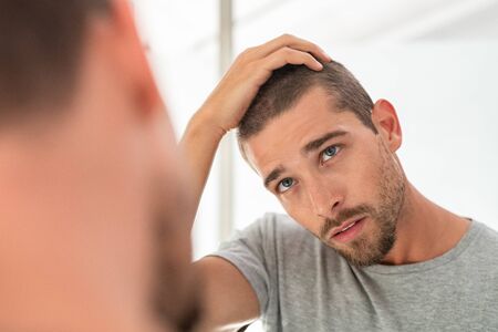 Young unshaven man looking at mirror in bathroom at home. Handsome guy looking at his face in mirror, checking hair and hairline. Man in pijamas concerned with hair loss.の写真素材