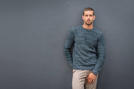 Handsome young cool man standing on grey background and looking at camera. Portrait of thoughtful casual man leaning against wall.Â Pensive stylish guy wearing sweater leaning against background with copy space.の写真素材