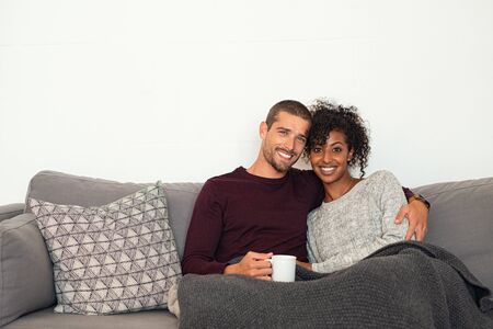 Happy multiethnic couple sitting on sofa with warm blanket, enjoying hot cup of tea. Young man and african woman embracing on couch in winter and looking at camera. Portrait of young couple in love hugging on sofa with copy space.の写真素材