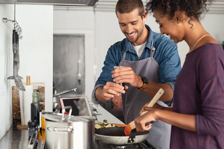 Young man cooking with girlfriend and adding spice to the sauce. Guy adds black pepper into frying pan on stove while woman using spatula to mix. Multiethnic couple preparing lunch together at home.の写真素材