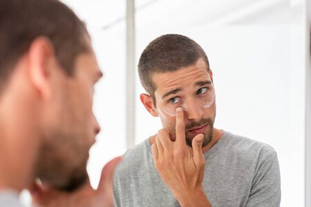 Young man taking care of his undereye wrinkles putting anti aging eye moisturizer. Man applying skincare facial treatment cream on face in the bathroom. Handsome guy applying moisturizer and looking at himself while standing in front of the mirror.