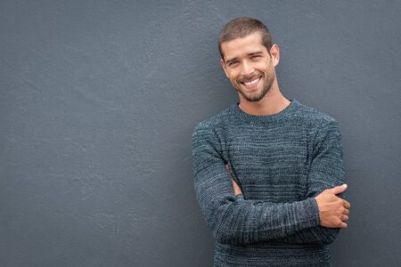 Portrait of happy young man leaning against wall isolated on grey background with a big smile. Handsome cheerful guy in winter clothes on gray wall looking at camera. Stylish man wearing sweater with crossed arms standing against wall with copy space.の写真素材