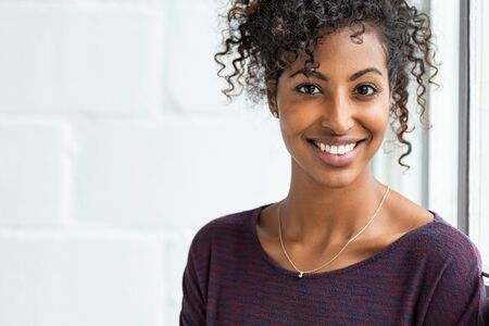 Portrait of a beautiful african woman smiling. Young black woman in casual looking at camera with copy space. Portrait of cheerful girl with afro hair sitting near a window.の写真素材
