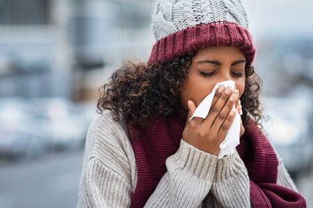 Pretty black woman blowing her nose with a tissue outdoor in winter. Young african woman getting sick with flu in a winter day. Woman with a cold rubs nose with handkerchief.の写真素材