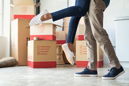 Man lifting his girlfriend among cardboard boxes as they start living together in new house. Closeup of excited husband embracing wife celebrating relocation. Loving man standing near boxes lifting woman up in the air excited about moving in.の写真素材