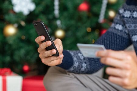 Closeup of man hand holding smartphone and credit card making payment during christmas time. Man in blue winter sweater paying online near xmas tree and buying gift. Man doing shopping online with debit card and mobile phone.の写真素材