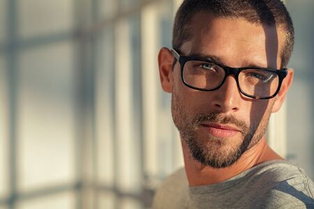 Portrait of stylish young man wearing eyeglasses at home. Closeup face of handsome guy wearing spectacles with sunlight and shadow reflection on face. Portrait of satisfied cool man in casual looking at camera with copy space at sunset.の写真素材