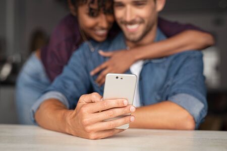 Happy couple using smartphone at home. Closeup of smiling man holding smartphone while african woman embracing him in background. Couple sharing media on smart phone.の写真素材