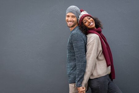 Young multiethnic couple holding hands while standing and leaning on each other back. Happy man and black woman standing back to back and smiling against grey wall with copy space. Cheerful guy and beautiful woman in warm clothes looking at camera isolated.の写真素材