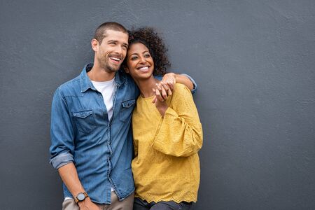 Young multiethnic couple in love isolated on grey background looking up and thinking about their future together. Smiling man and african woman in casual hugging and looking away while planning the future. Cheerful couple holding hands while leaning on wall.の写真素材