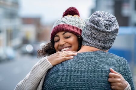 Happy african american girlfriend hugging boyfriend on street with closed eyes. Friends meeting and embracing outdoor. Romantic multiethnic young couple embracing in winter clothes in the city.の写真素材