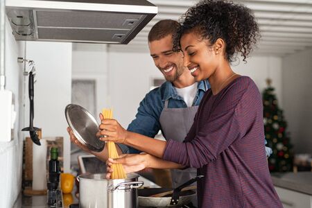 Smiling woman adding pasta to the pot while man holding lid. Happy couple adding spaghetti to boiling water while preparing tasty Italian cuisine. Cheerful couple preparing pasta in pot for dinner in kitchen.の写真素材