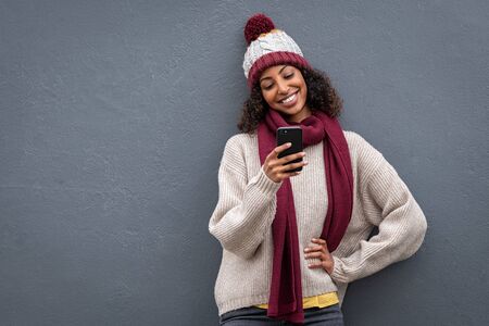 Young african woman using mobile phone standing against grey wall. Smiling beautiful girl wearing warm clothes sending message from cellphone. Black woman wearing sweater and knitted hat while surfing the net with smartphone isolated on gray background with copy space.の写真素材