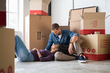 Cheerful couple in conversation in new home with cardboard boxes around. Romantic african girl lying down with head on boyfriend lap while sitting on floor with unpacked boxes in new apartment. Newly married couple excited looking at each others..の写真素材
