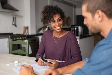 Young multiethnic couple doing accounting online with a phone bank app. Portrait of african casual girl sitting on table at home and analyzing home finance with documents. Girl showing the current account on the mobile app to her boyfriend.の写真素材