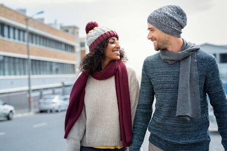 Casual young couple holding hands while walking on the street. Happy couple wearing winter clothes and looking at each other. Romantic multiethnic man and woman enjoying winter breeze outdoor.の写真素材