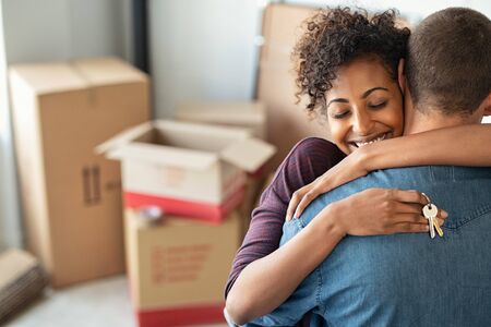 Young african woman holding home keys while hugging boyfriend in their new apartment after buying real estate. Lovely girl holding keys from new home and embracing man. Happy couple in their apartment around cardboard boxes.の写真素材