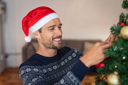 Happy man decorating christmas tree at home with santa claus hat. Handsome guy wearing xmas sweater decorating christmas tree. Man standing and decorating tree with golden baubles during winter holiday.の写真素材