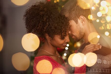 Romantic couple in sweaters dancing before new year eve at home. Young lovely man and african woman hugging over christmas bokeh lights. Multiethnic couple dancing in love with golden lights bokeh in background.