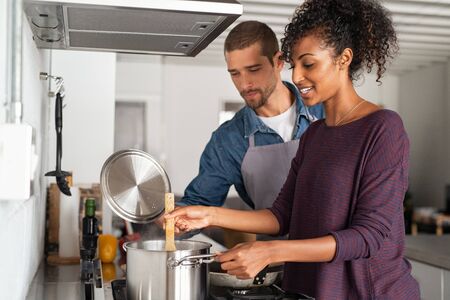 Portrait of happy young couple cooking together in kitchen at home. Woman cooking in pot with her boyfriend in kitchen. Man in apron helping his girlfriend preparing dinner.の写真素材