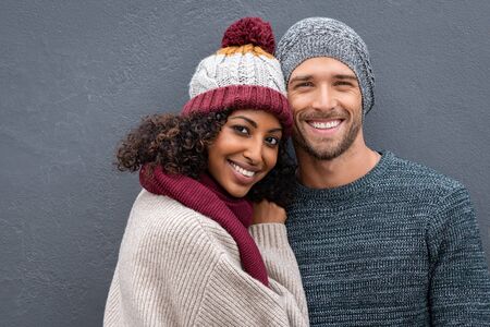 Young multiethnic couple in warm clothing looking at camera on a winter day isolated on gray wall with copy space. Portrait of smiling man and beautiful happy woman wearing sweater and woolen cap, hugging isolated against grey background.の写真素材