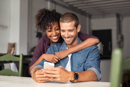 Smiling young couple embracing while looking at smartphone. Multiethnic couple sharing social media on smart phones while sitting at table. Smiling african girl embracing from behind her happy boyfriend while using cellphone and laughing.の写真素材