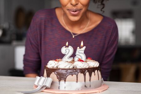Happy young woman blowing birthday candle at home. Closeup of delicious cake with cream and chocolate on table while woman making birthday wish. Cheerful girl celebrating her 25th birthday at home.の写真素材
