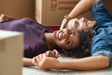 Happy multiethnic couple smiling while lying on floor near cardboard boxes. Portrait of african woman looking at camera while cheerful man lying on the floor of their new home. Young casual couple moving house.の写真素材
