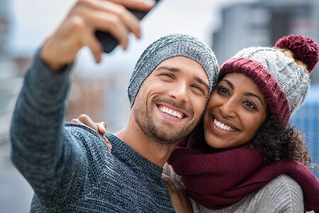 Happy multiethnic couple in the city taking selfie in a winter day. Smiling man and african girl taking selfie with smartphone during winter on street. Young woman and cheerful guy on christmas holidays taking photo.の写真素材