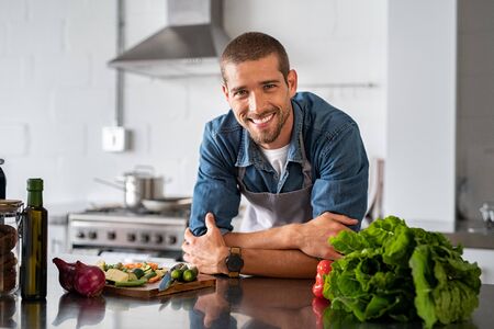Handsome young man leaning on kitchen counter with vegetables and looking at camera. Smiling man wearing apron while preparing food at home. Portrait of happy casual guy leaning on steel counter in the kitchen with vegetables and ingredients on it.の写真素材