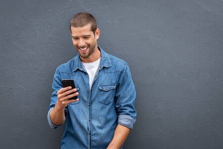 Young man leaning against a grey wall using mobile phone with copy space. Happy casual guy messaging on smartphone on gray background. Cheerful man looking down while typing and reading a message on cell phone.の写真素材