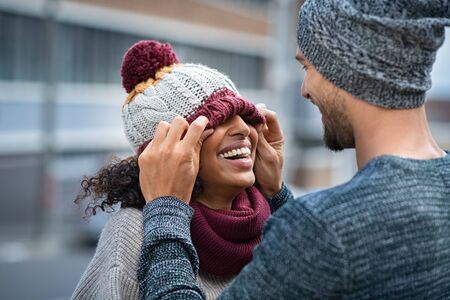 Loving multiethnic couple having fun outdoor in winter. Young man covering eyes his girl with woolen cap. Cheerful guy and smiling african woman playing in winter outdoor.の写真素材