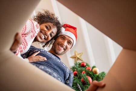 Happy multiethnic couple embracing while looking inside a gift box. Young man and beautiful african woman unpacking xmas present with christmas tree. Smiling young man wearing santa claus hat with woman looking at camera while opening present.の写真素材