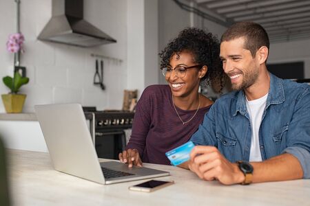 Smiling young couple making shopping online with credit card and laptop at home. Happy multiethnic couple holding debit card while buying on ecommerce site using laptop. Cheerful guy and african girl making online purchase.の写真素材